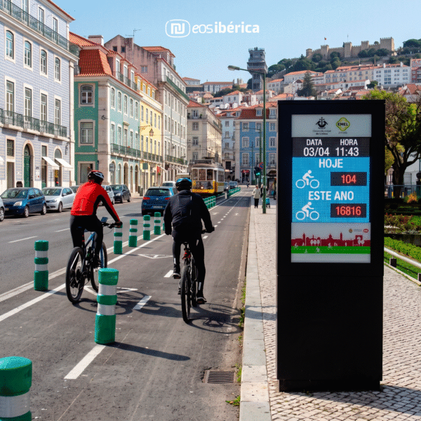 Dois ciclistas numa ciclovia em Lisboa ao lado de um tótem digital que exibe o número de bicicletas contadas 'Hoje' e 'Este ano'.